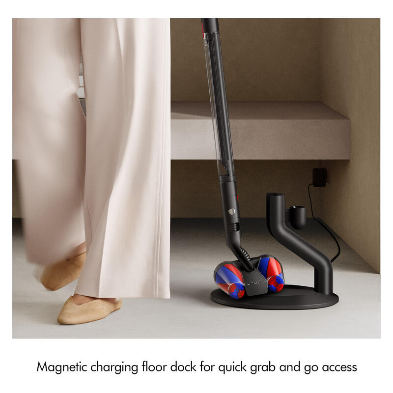 A cordless vacuum cleaner with a red and blue brush head is shown charging in a black magnetic floor dock beside a person wearing beige pants and flats in a minimalist indoor setting. Text at the bottom reads: 'Magnetic charging floor dock for quick grab and go access'.