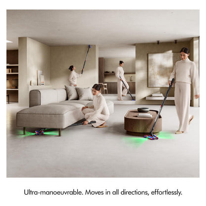 Three women in matching cream loungewear clean a modern open-plan apartment with a long-handled floor mop and a small floor sweeper, with green floor light accents. The scene includes a gray sectional sofa, a round wooden coffee table, and a large abstract painting on the wall.