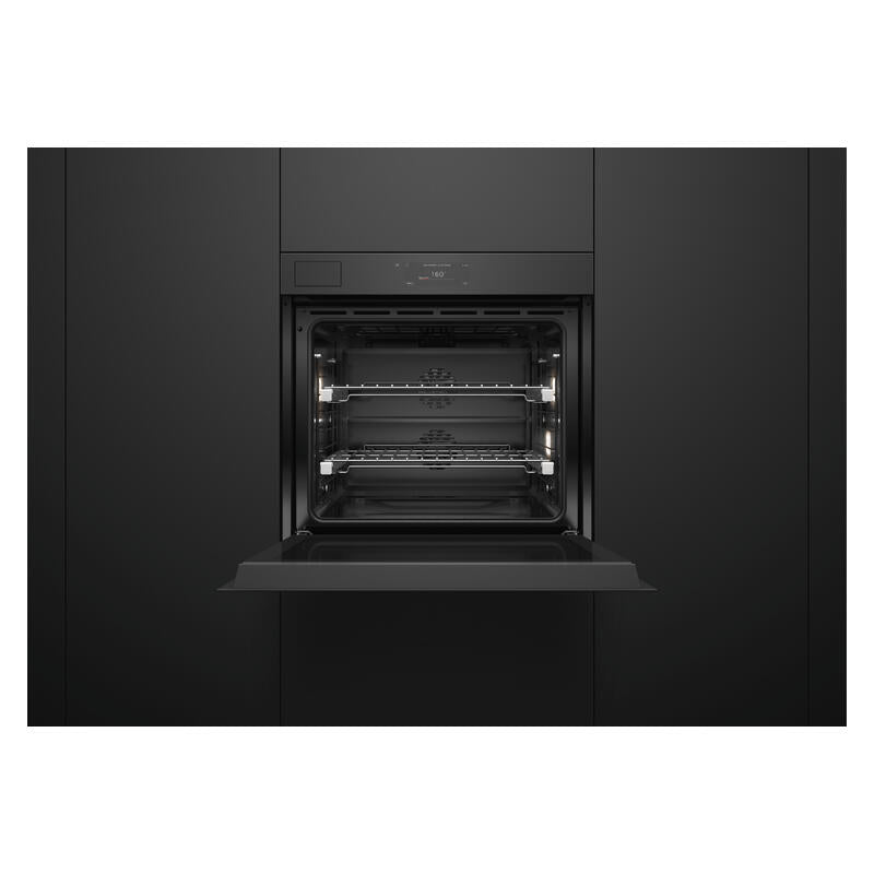 A built-in wall oven shown from the front with its door open, revealing two interior racks and lit oven lights, set against a dark charcoal kitchen cabinet background.