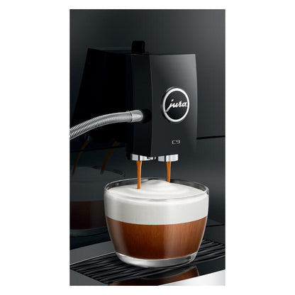 A black Jura C9 espresso machine dispensing espresso into a glass cup filled with milk and foam, shown against a dark studio background.