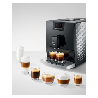 A Jura coffee machine in black and silver, shown dispensing espresso and latte drinks into small glasses on a light gray studio background.