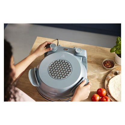 A person uses a countertop electric pizza oven on a wooden table, with dough, tomatoes, and basil visible around it.