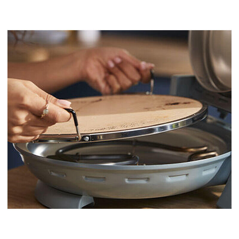 Close-up of a person's hands lifting a round wooden lid with metal handles off a shallow round cooking pan or grill base.