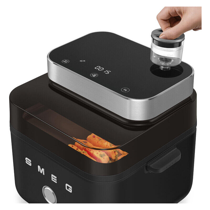 A black and silver SMEG electric food air fryer with a clear lid, shown with a hand placing a small clear container into the top opening. A glass window on the front reveals cooked food inside.
