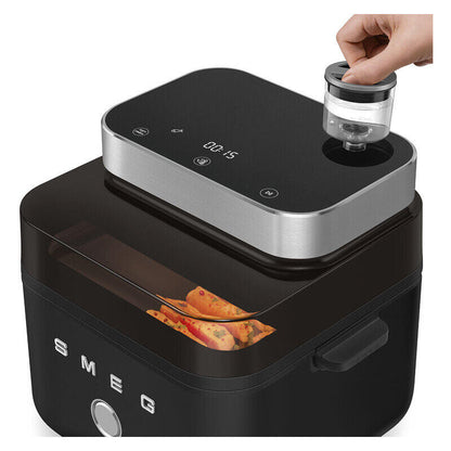 A black and silver SMEG electric food air fryer with a clear lid, shown with a hand placing a small clear container into the top opening. A glass window on the front reveals cooked food inside.